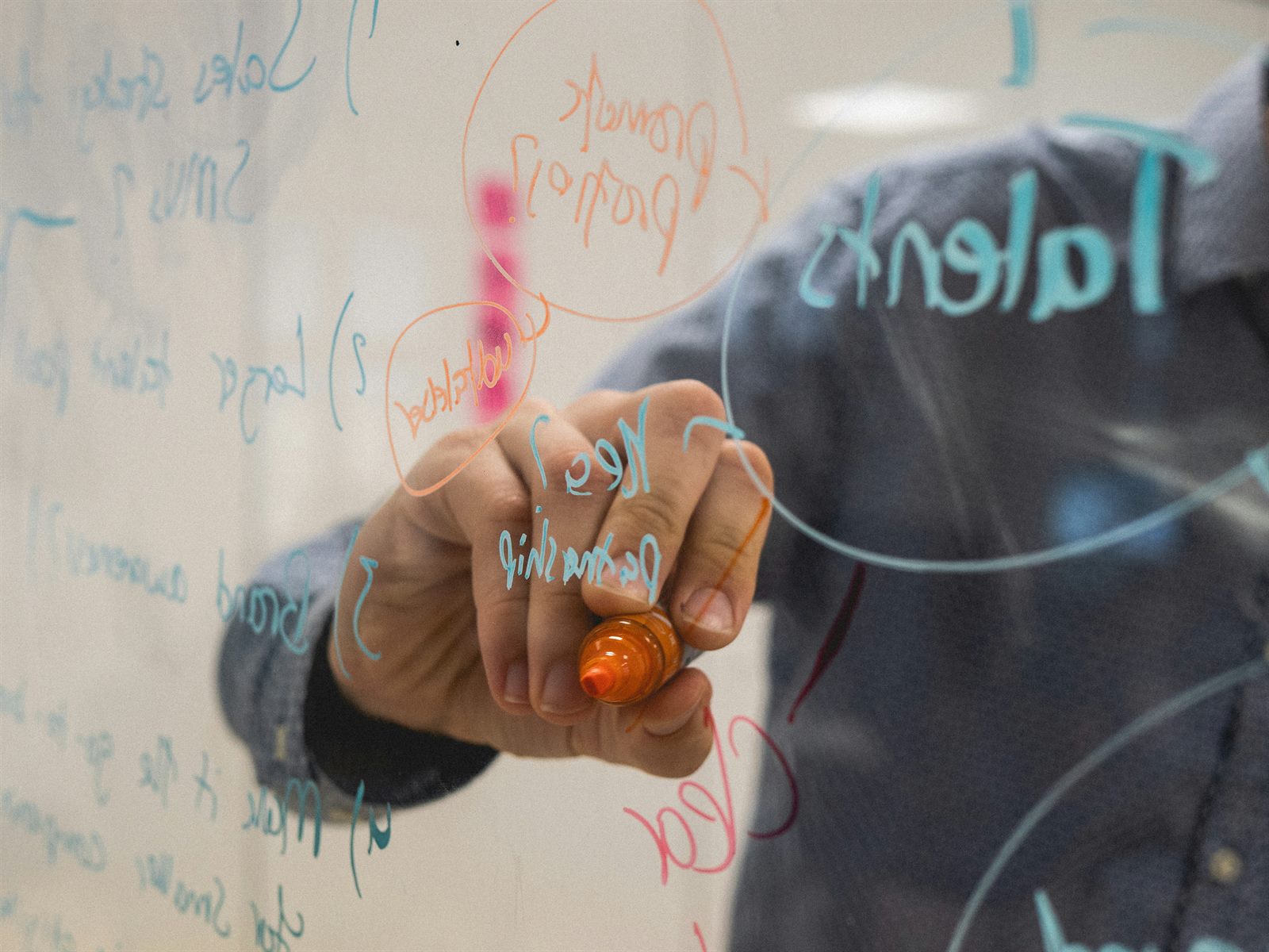 A young person sketching ideas on a whiteboard in a modern open-plan office, laptop closed beside them