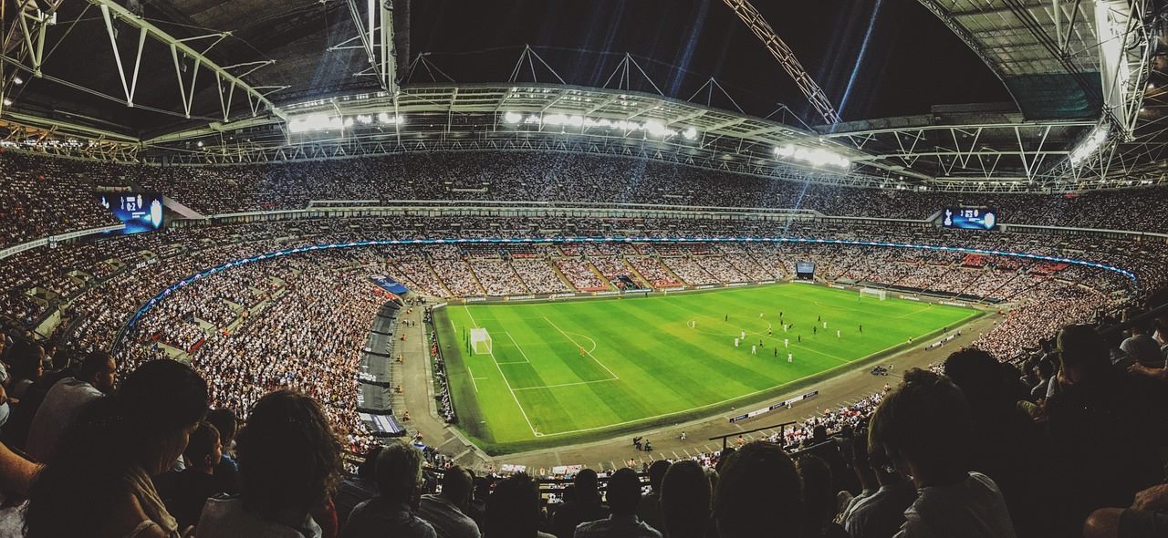 Football fans in a stadium reacting with astonishment to a record transfer figure displayed on the big screen
