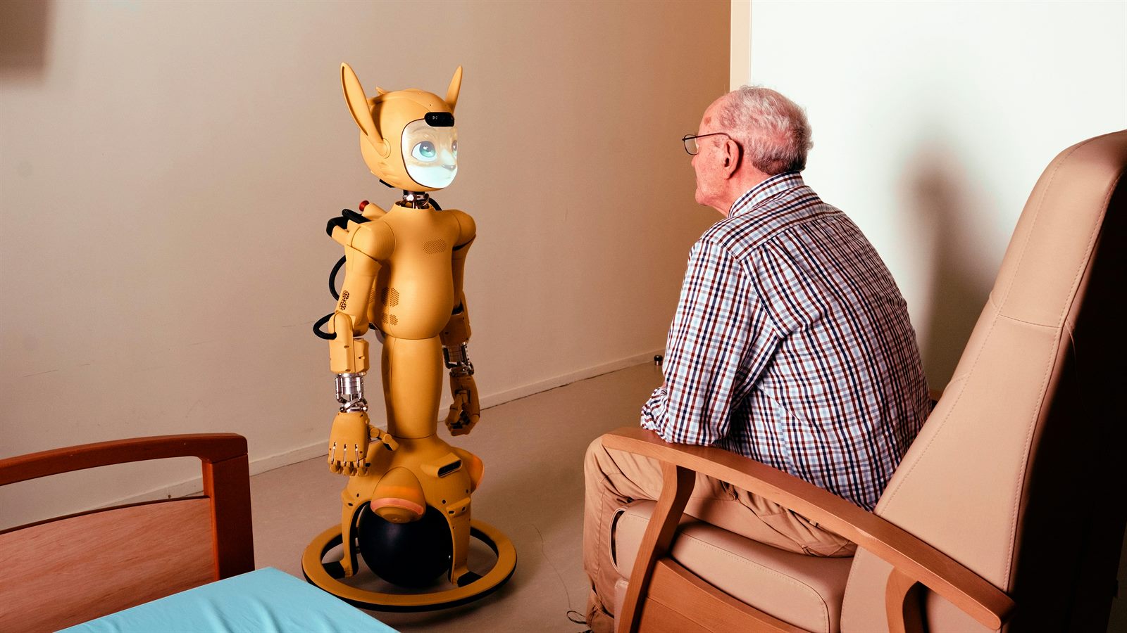 Elderly woman in a care home smiling at a small companion robot on the table beside her tea